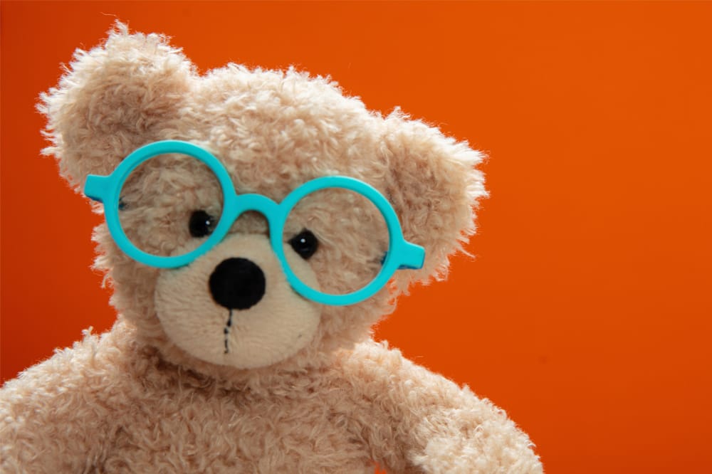 Image shows a teddy bear wearing glasses against an orange background.